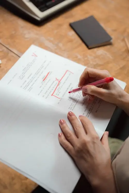 Auf diesem Bild ist die Hand einer Person zu sehen, die eine Stelle in einem gedruckten Text mit einem roten Stift markiert. Sie macht Kreuze, unterstreicht einzelne Passagen und macht Anmerkungen.